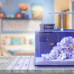 a small rimless saltwater aquarium with white sand and white rock inside. Two orange clownfish are swimming, and the tank has a hang on the back filter and small led light attached. The aquarium is sitting on a grey wood table with a blurred bright bedroom in the background